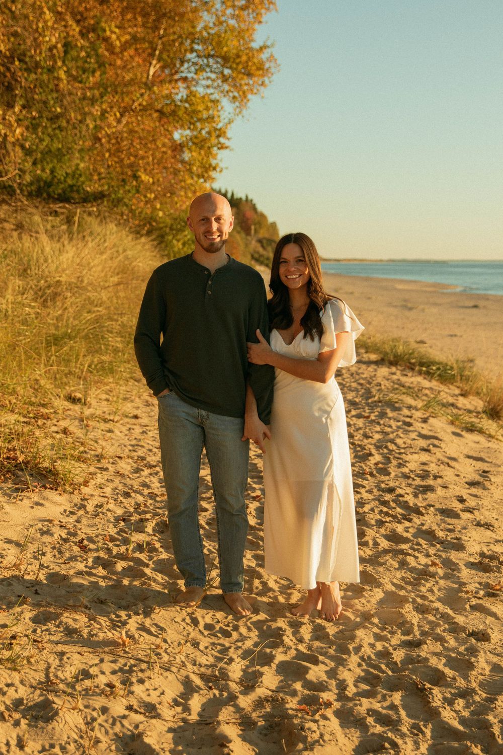 Smiling couple standing barefoot on a sandy beach path surrounded by autumn foliage at golden hour.
