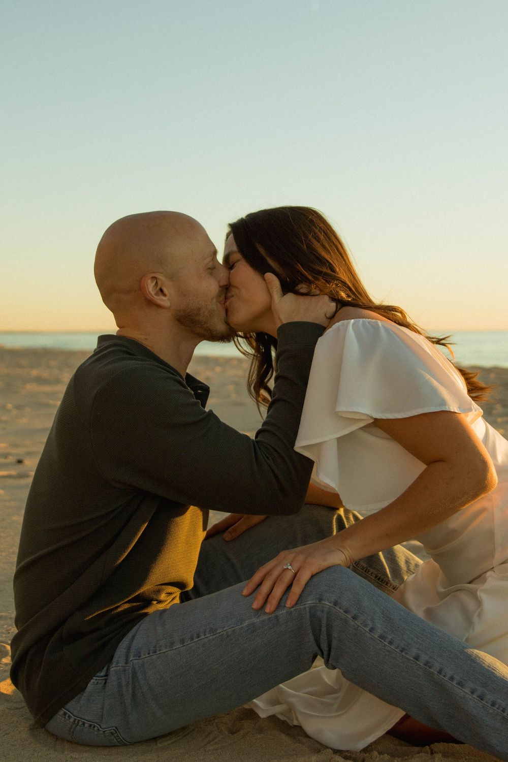 Couple sharing a romantic kiss while sitting on a sandy beach at sunset.