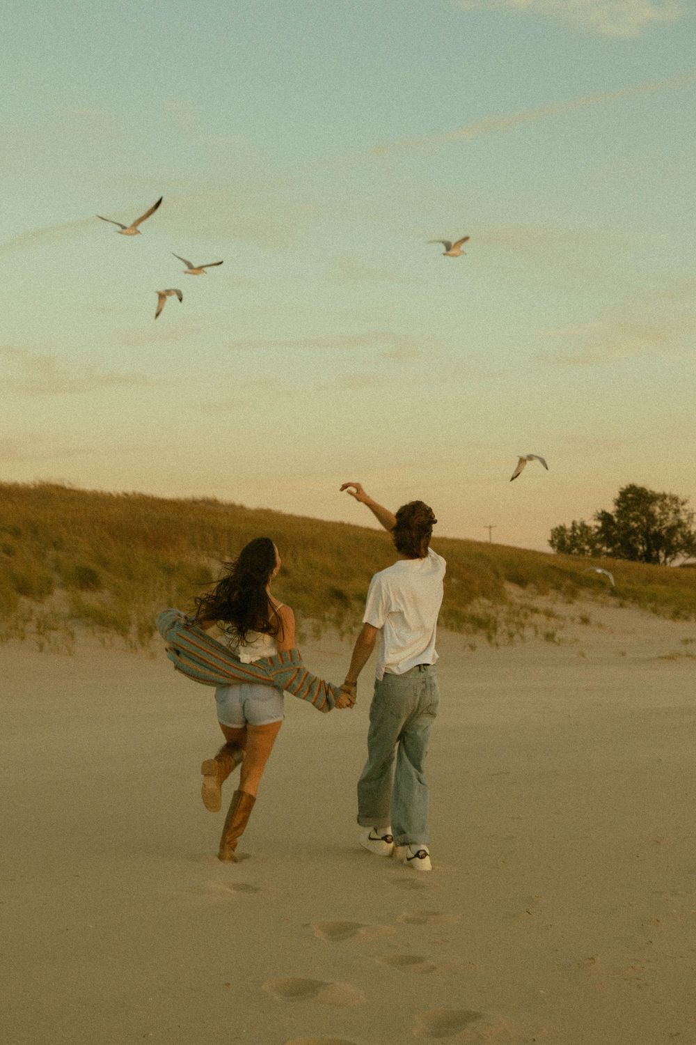 Couple holding hands on a beach at dusk with seagulls flying overhead.