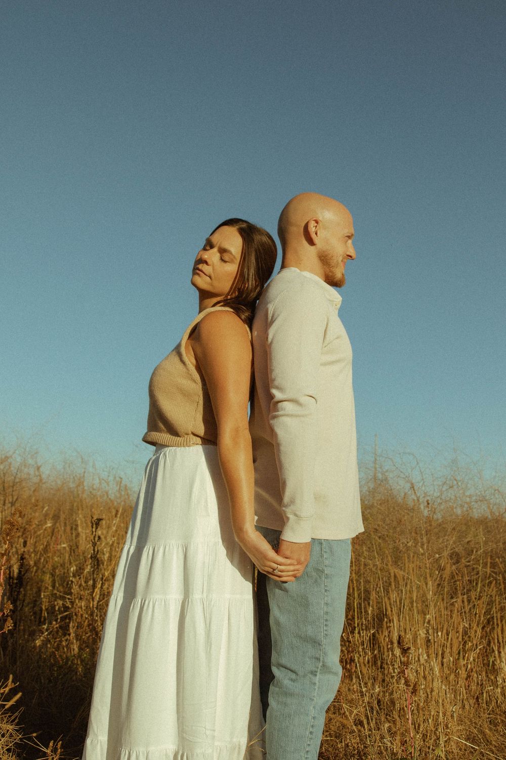 Couple standing back to back in a golden wheat field under a clear blue sky.