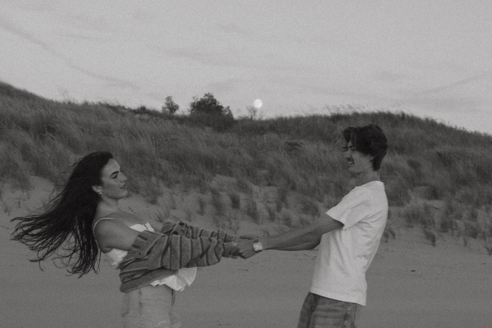 Black and white photo of a couple playfully spinning on a sandy beach dune.