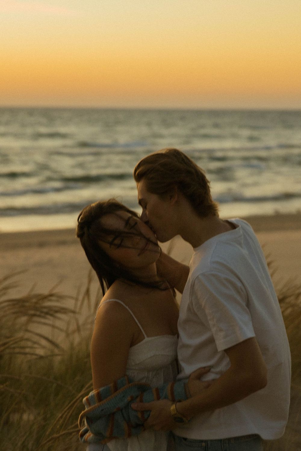 Couple sharing a romantic kiss on a sandy beach at golden sunset with ocean waves in the background.