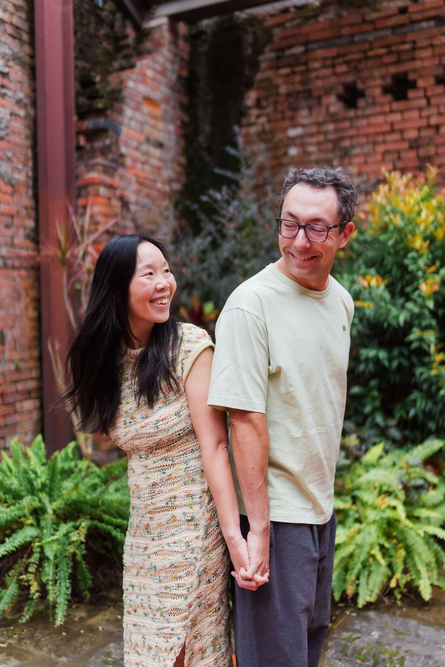 A couple shares a joyful moment together in front of a rustic brick wall with lush green ferns at Bopiliao Historical Block in Taiwan.