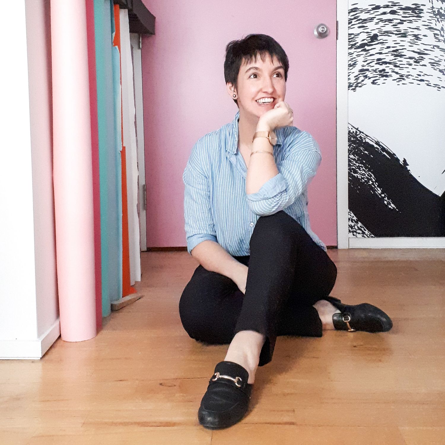 Person in blue shirt and black pants sitting casually on wooden floor against pink wall.