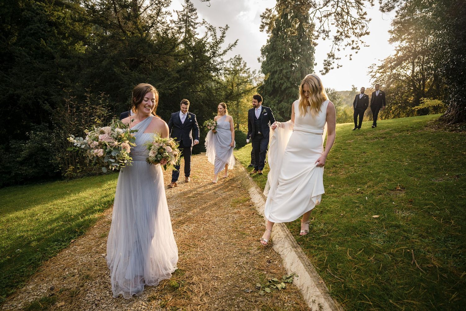 Wedding party walking along a scenic dirt path through green gardens during golden hour lighting.