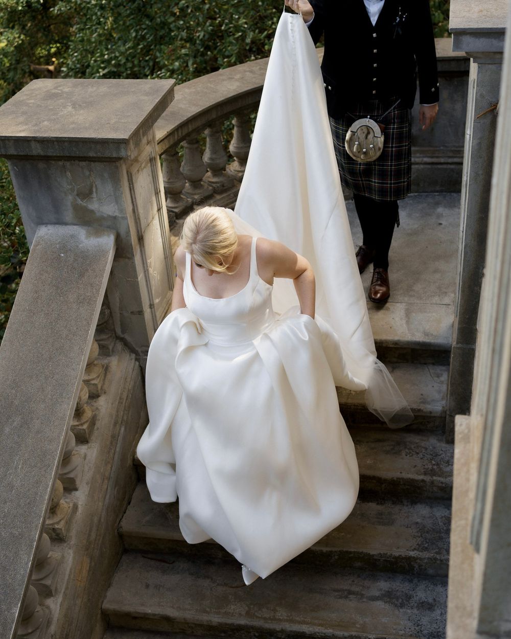 Bride in white gown ascending stone steps with person in traditional kilt behind.