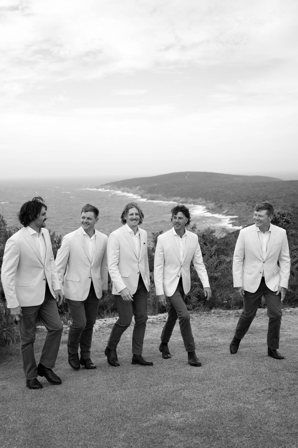 Five groomsmen in white jackets posing casually on a hilltop overlooking the ocean.