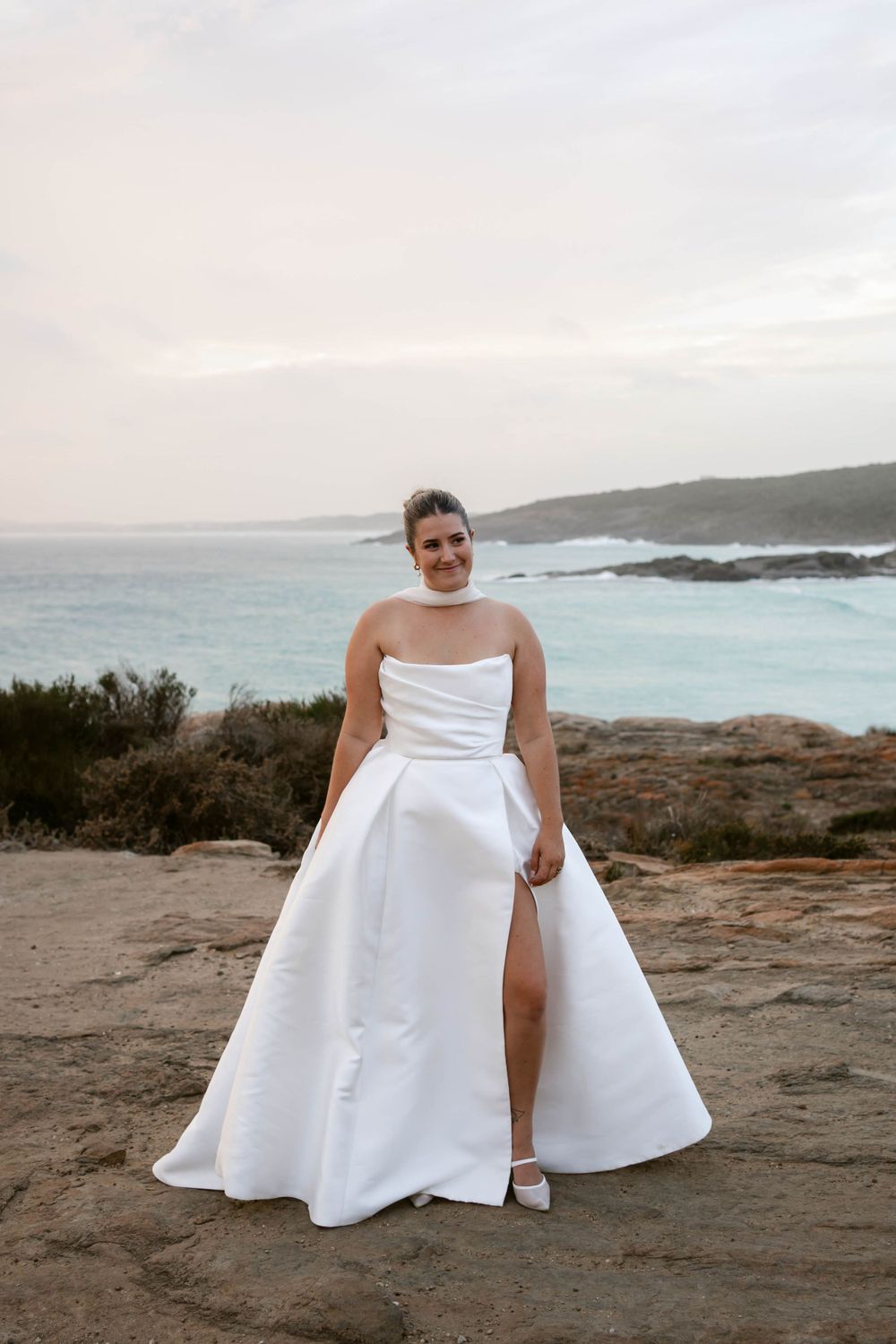 A bride in a white strapless wedding gown with a thigh-high slit poses on a coastal cliff at sunset.