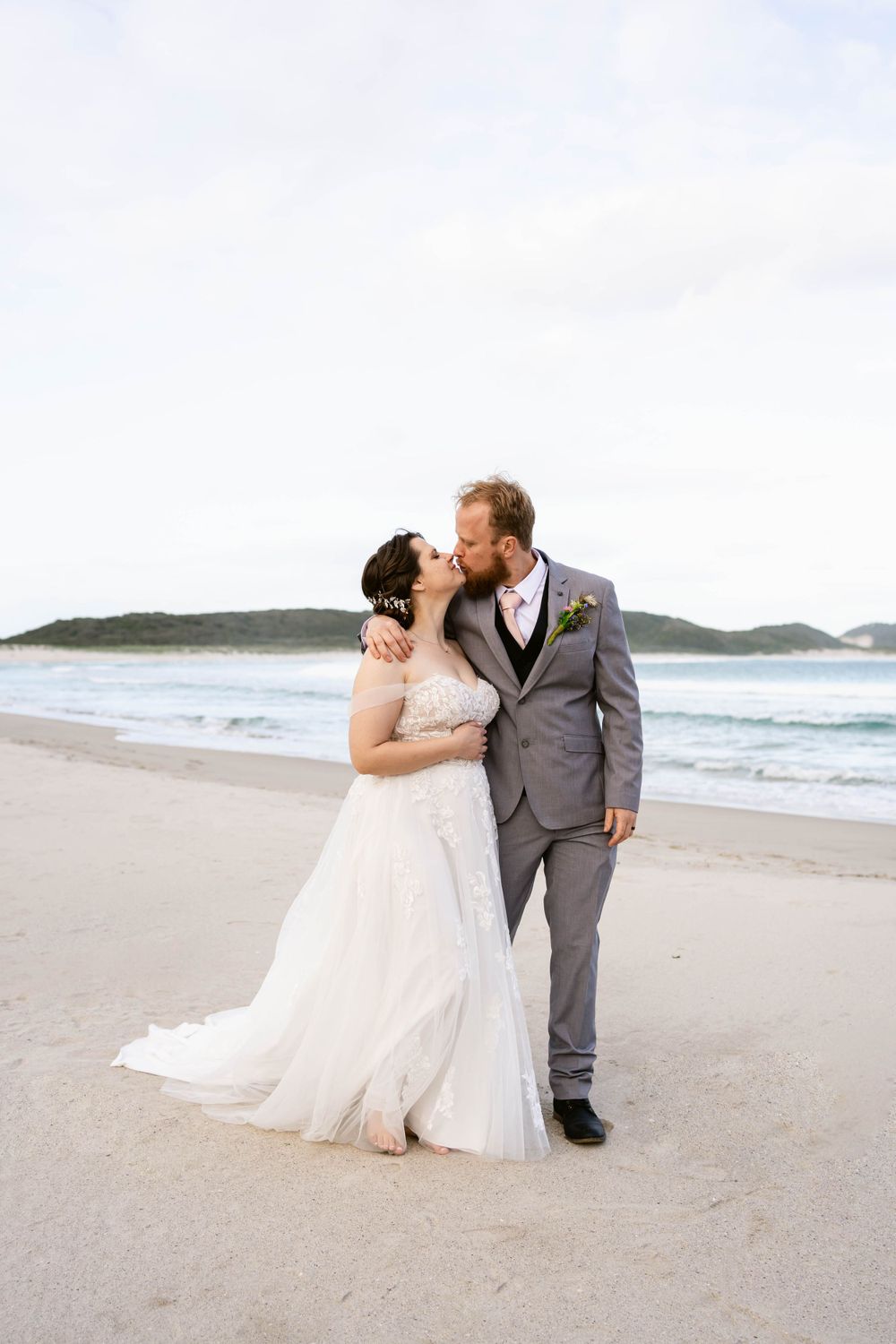 Romantic beachside wedding portrait with ocean waves and cloudy sky in background.