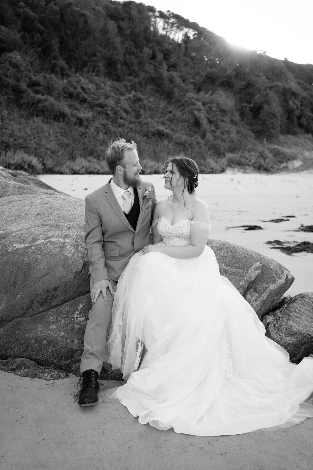 Black and white wedding photo of couple sitting on coastal rocks at sunset.