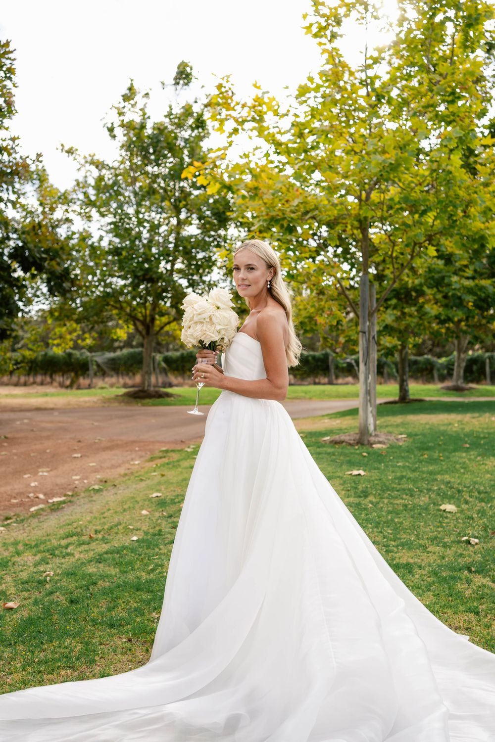 Bride in a stunning white ballgown with long train poses with bouquet in an outdoor garden setting.