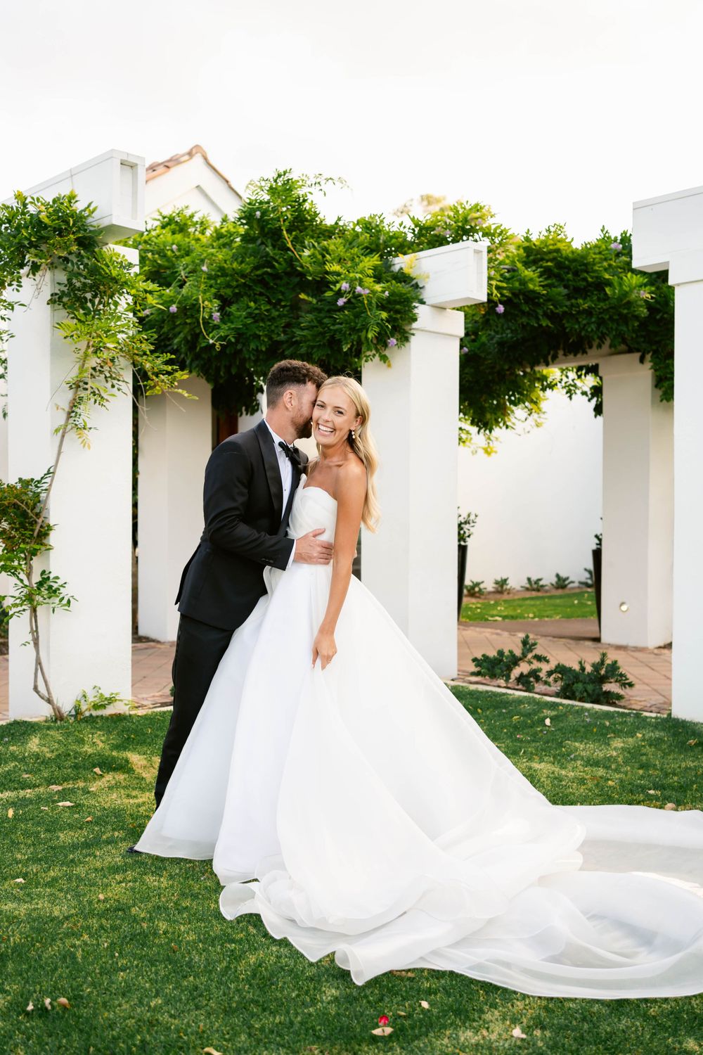 Elegant wedding portrait of a couple in formal attire standing by white pillars adorned with green vines.