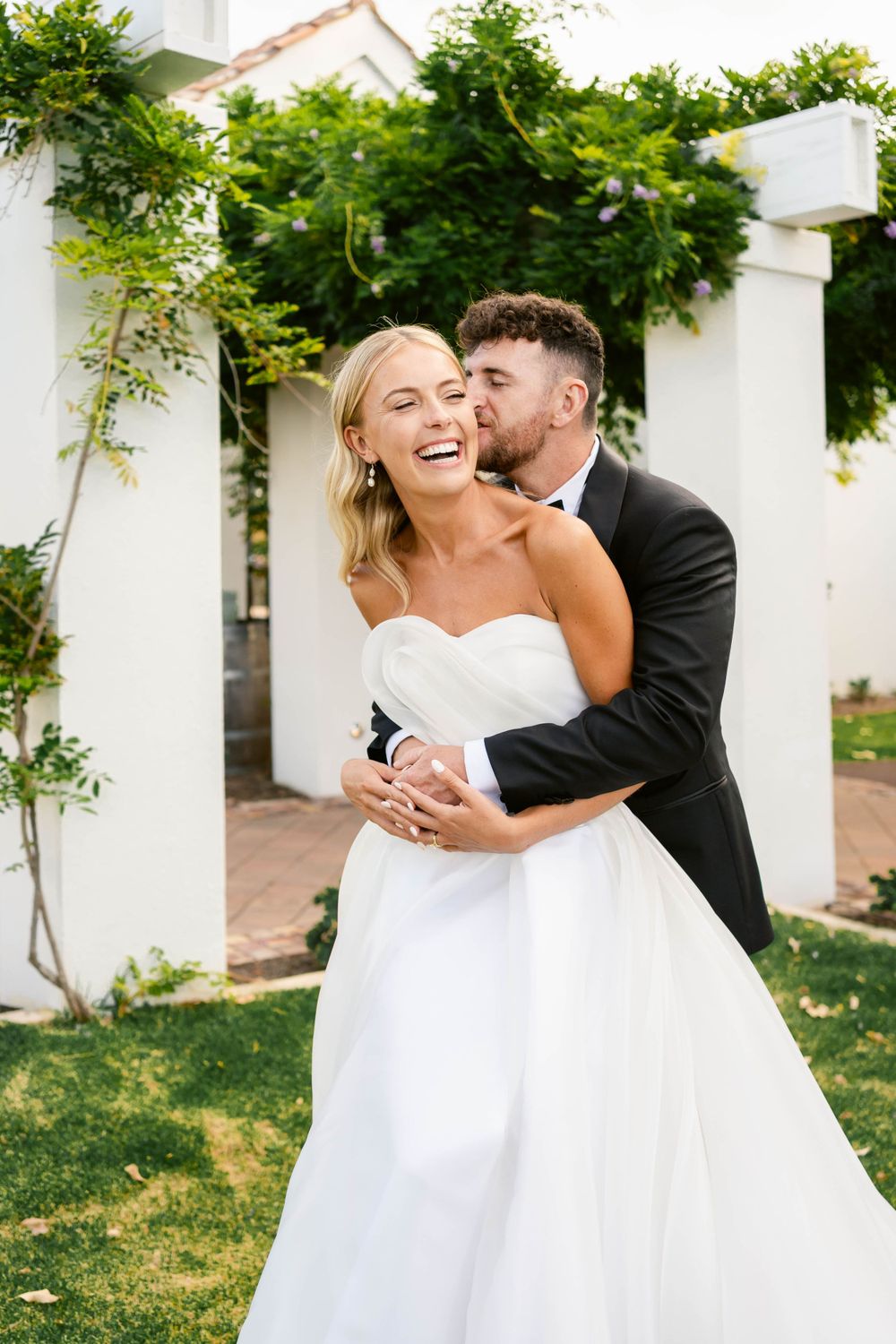A joyful wedding moment captured outdoors with a white strapless gown and classic black tuxedo against a vine-covered backdrop.