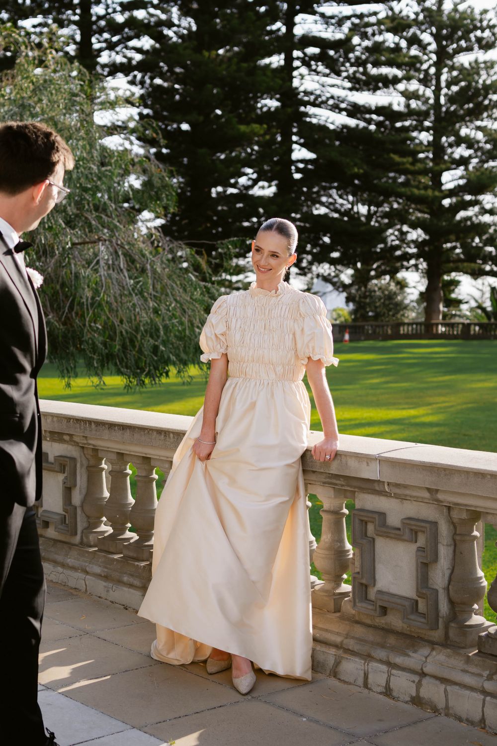 A couple in formal wedding attire standing on an elegant stone balcony with pine trees in background.