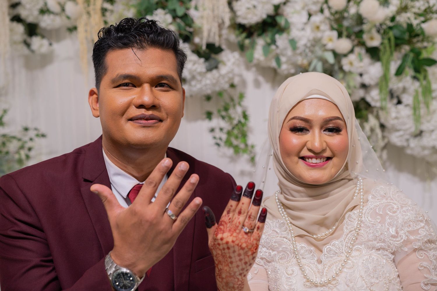 A wedding couple in burgundy and white attire showing their rings against a floral backdrop.