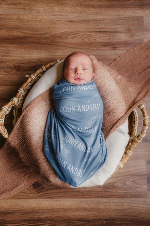 A newborn wrapped in a blue personalized swaddle blanket with 'JOHN ANDREW' text lies in a woven basket on wooden floor.