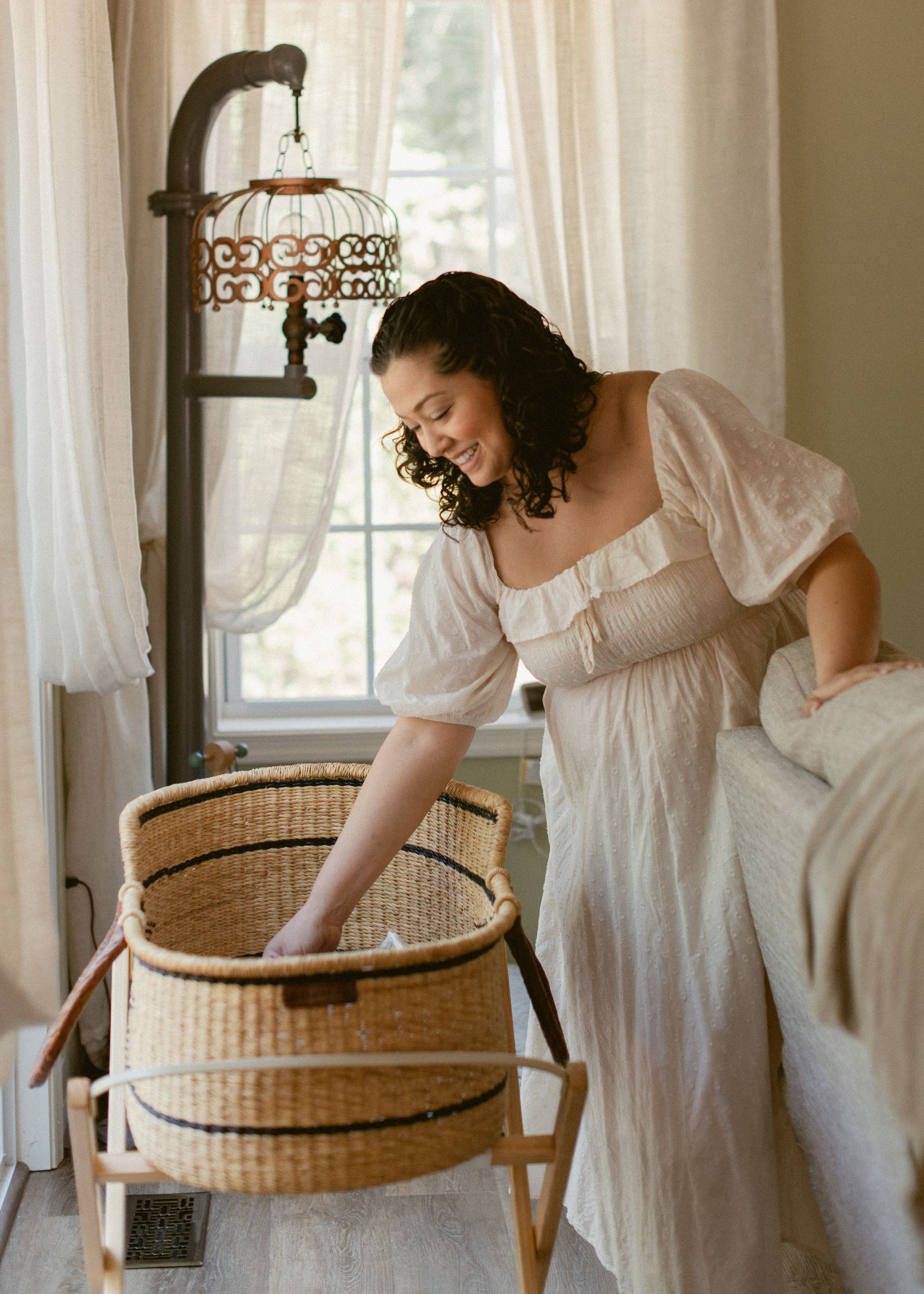 Woman in white vintage nightgown smiles while reaching into a woven basket by a window with an ornate lamp.