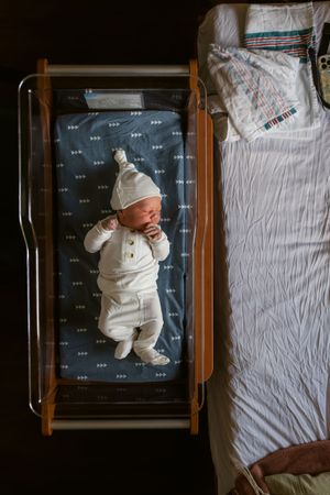 Newborn baby wearing white outfit and hat lies on dark blue patterned changing pad on wooden changing table.