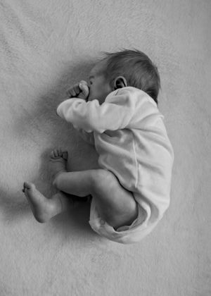 A tiny baby in a white onesie sleeps peacefully curled up in fetal position on a light gray blanket.