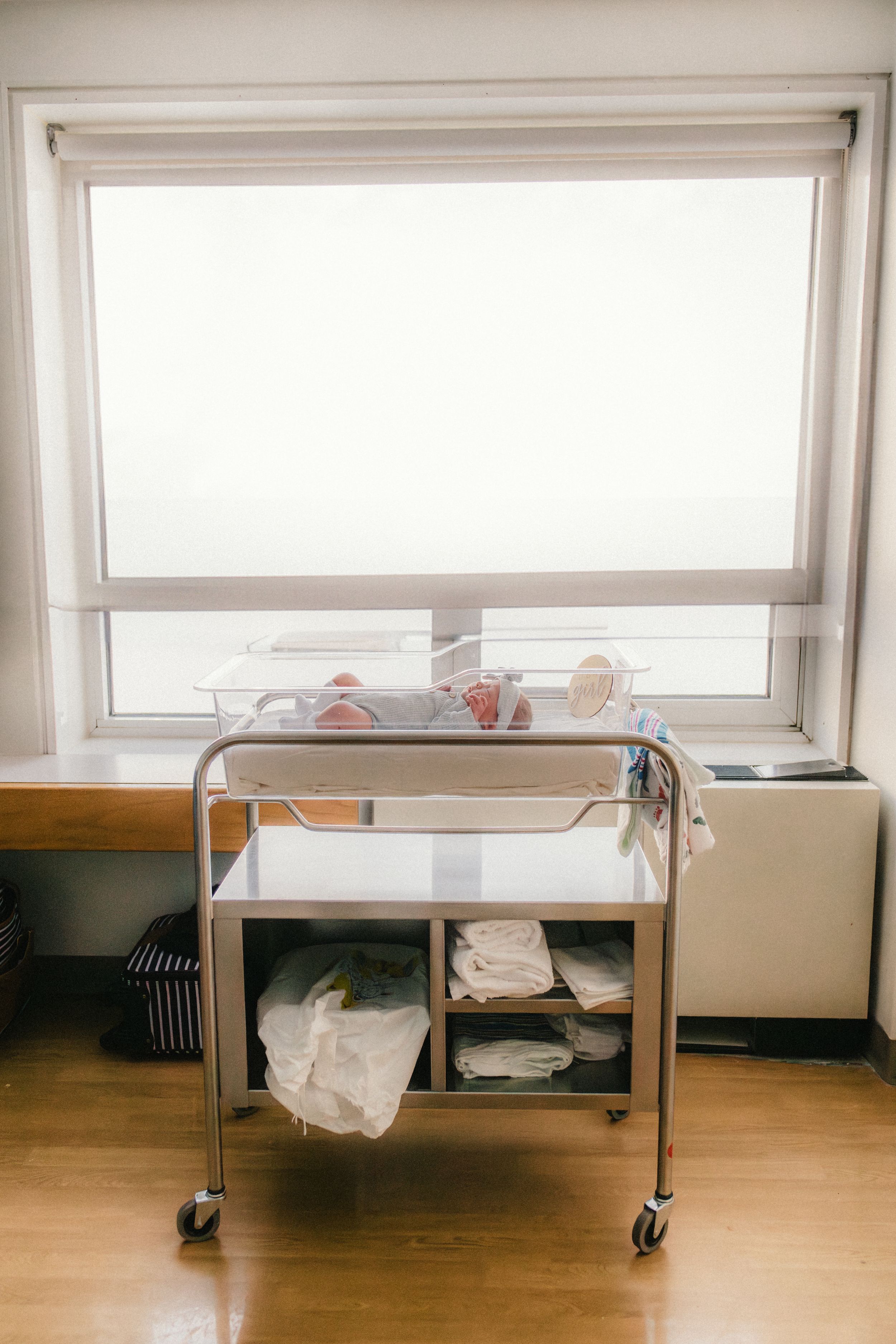 A hospital bassinet cart with supplies sits beneath a bright window.