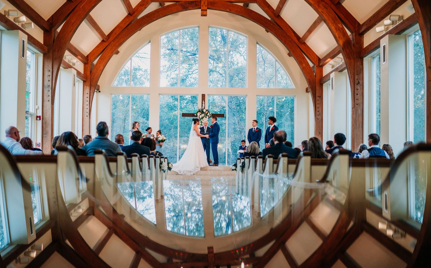 A wedding ceremony in an elegant chapel with a large arched window and wooden beams creating a mirror reflection on the floor.