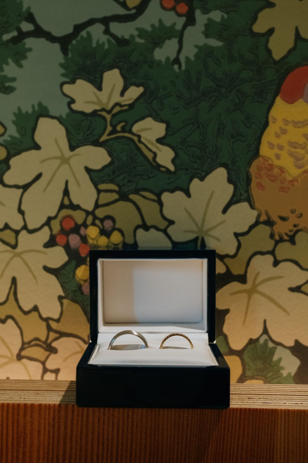 Wedding rings in a black box displayed against a vintage floral wallpaper background.