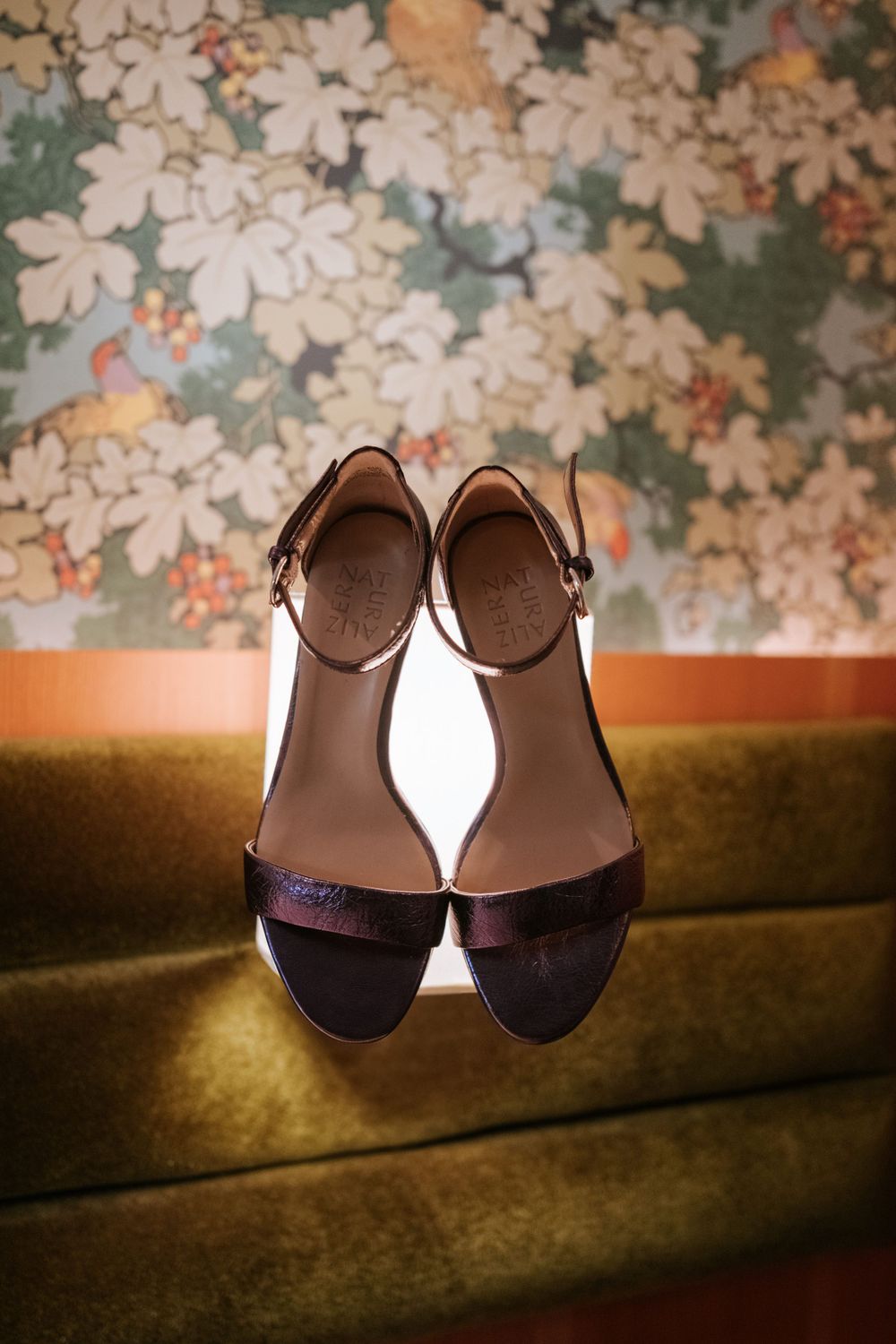 Elegant burgundy sandals resting on a gold velvet surface with floral wallpaper backdrop.