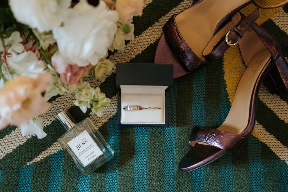 Wedding details including ring box, perfume bottle, and elegant burgundy heel on a striped green carpet.