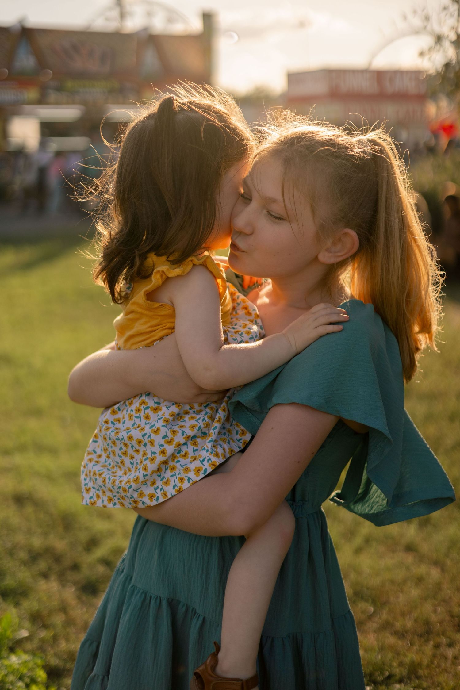 Young girl holds her sister at the fair in the sunset.