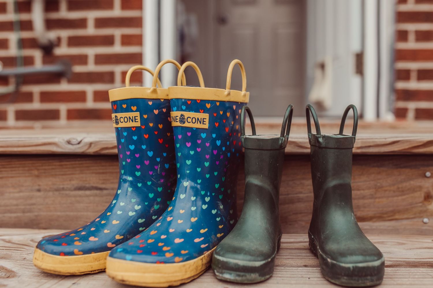 Two pairs of rubber rain boots, one blue with white polka dots and yellow trim, sit beside dark green boots on wooden steps. Whitney Popp