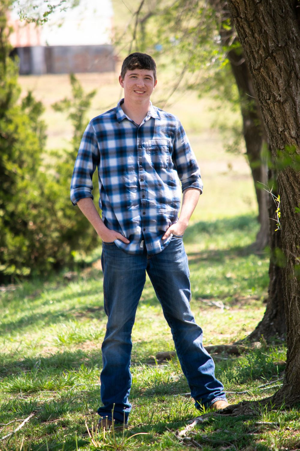 Smiling young man in a blue and gray plaid shirt and jeans, standing outdoors in a grassy area near trees.
