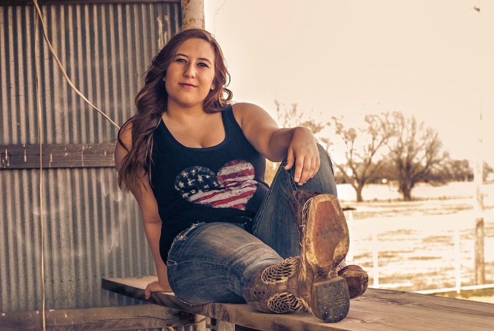 A woman in a heart-patterned tank top sits on a wooden beam, wearing jeans and cowboy boots, with a rural landscape in the background.