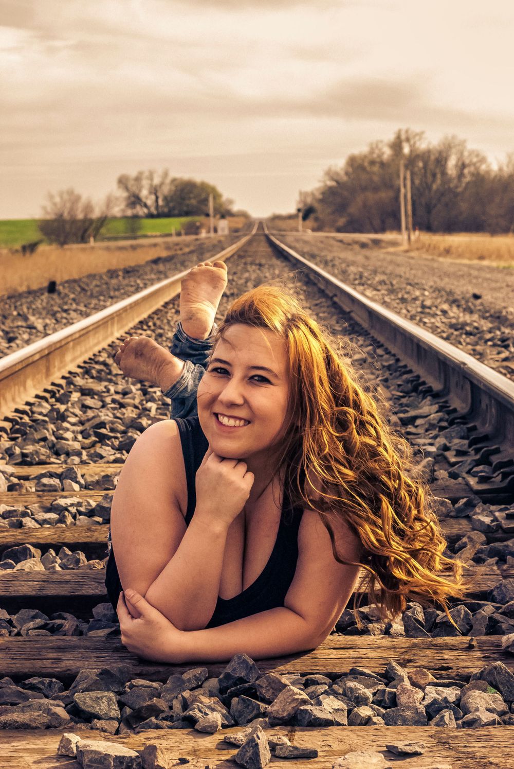 Woman with long auburn hair lying on railroad tracks, smiling, with bare feet and looking out at a distant horizon.