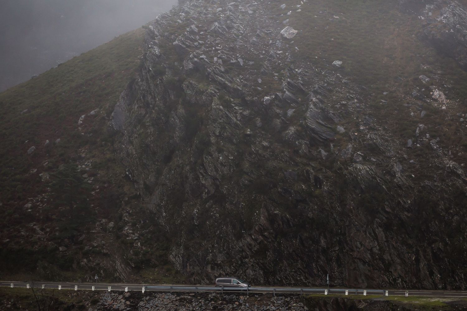A solitary car drives along a misty mountain road beside steep rocky cliffs in moody weather.