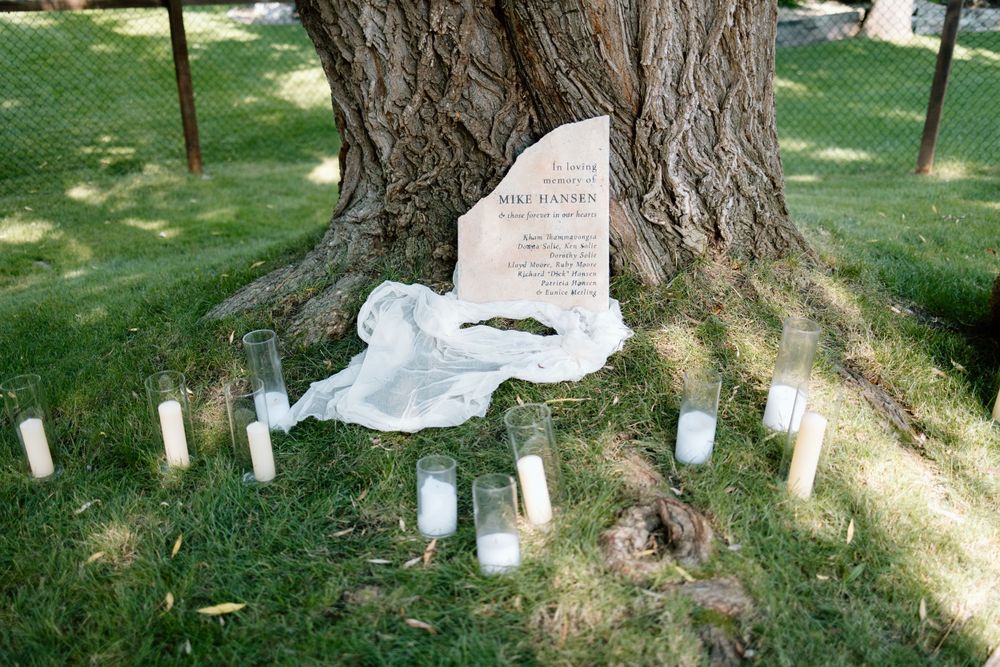 Memorial shrine with candles and white fabric arranged at base of large tree trunk with stone marker.