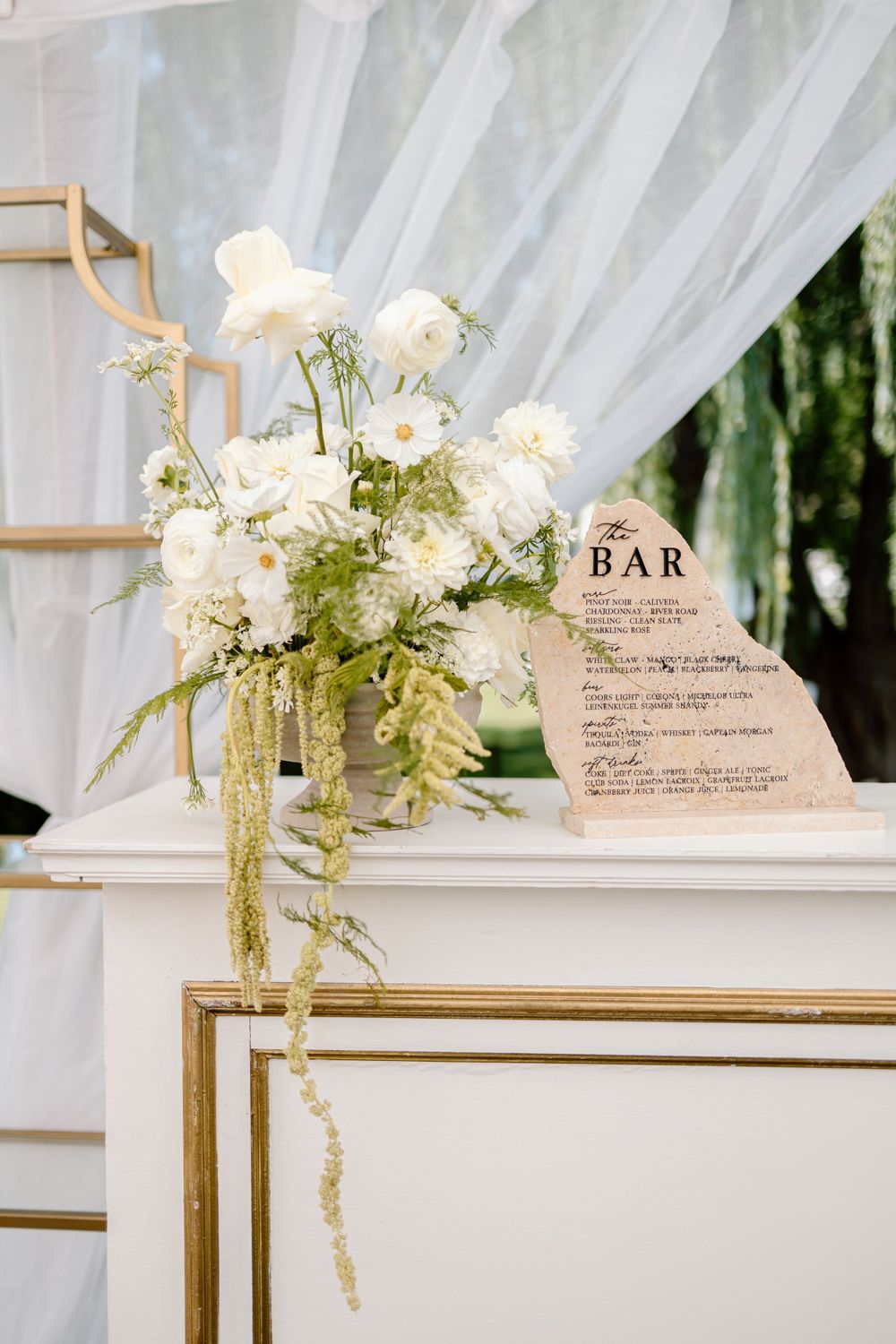 Elegant wedding bar menu display with white floral arrangement in gold frame and sheer drapery backdrop.