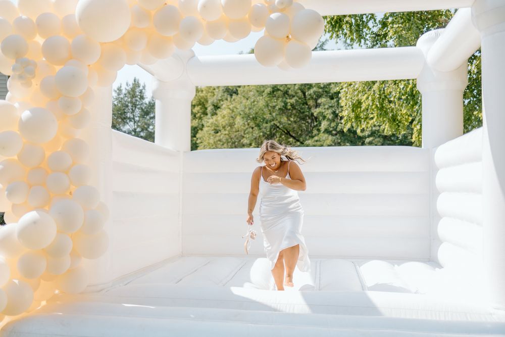 A person in a white dress jumps joyfully under a white archway decorated with string lights and greenery overhead.