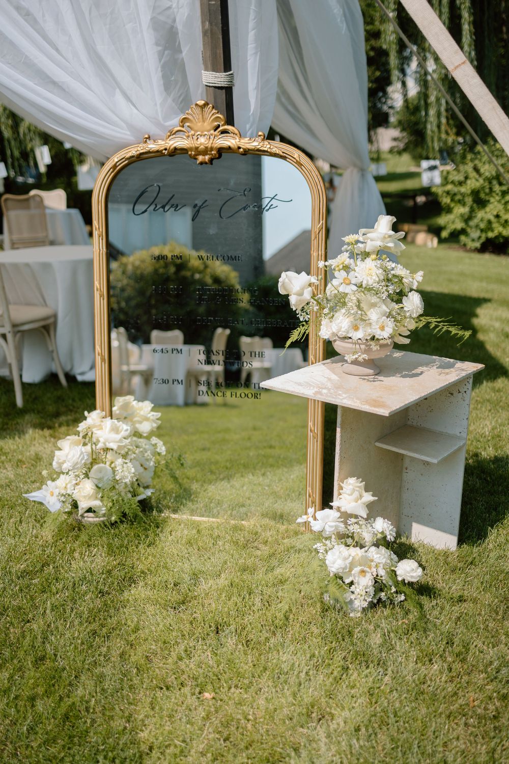 Elegant gold-framed mirror with white floral arrangements and sheer drapery creates a romantic outdoor wedding setting on grass.