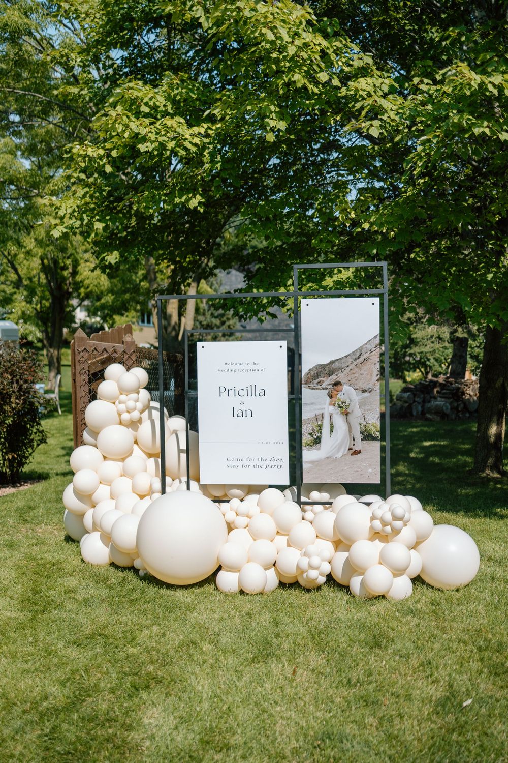 Wedding welcome sign display with white balloon arch and framed photos arranged on green lawn under leafy trees.