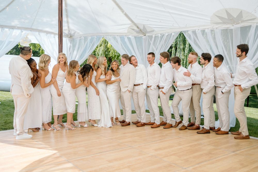 Wedding party dressed in all-white and khaki attire posing under a white tent.