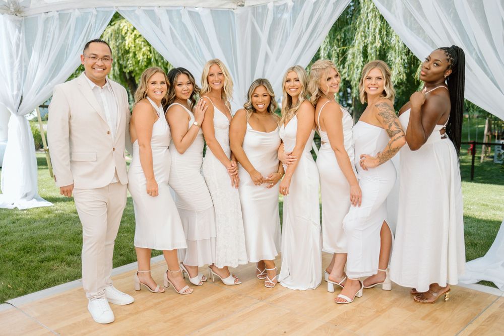 Wedding party in coordinated white outfits posing on outdoor dance floor under tent.