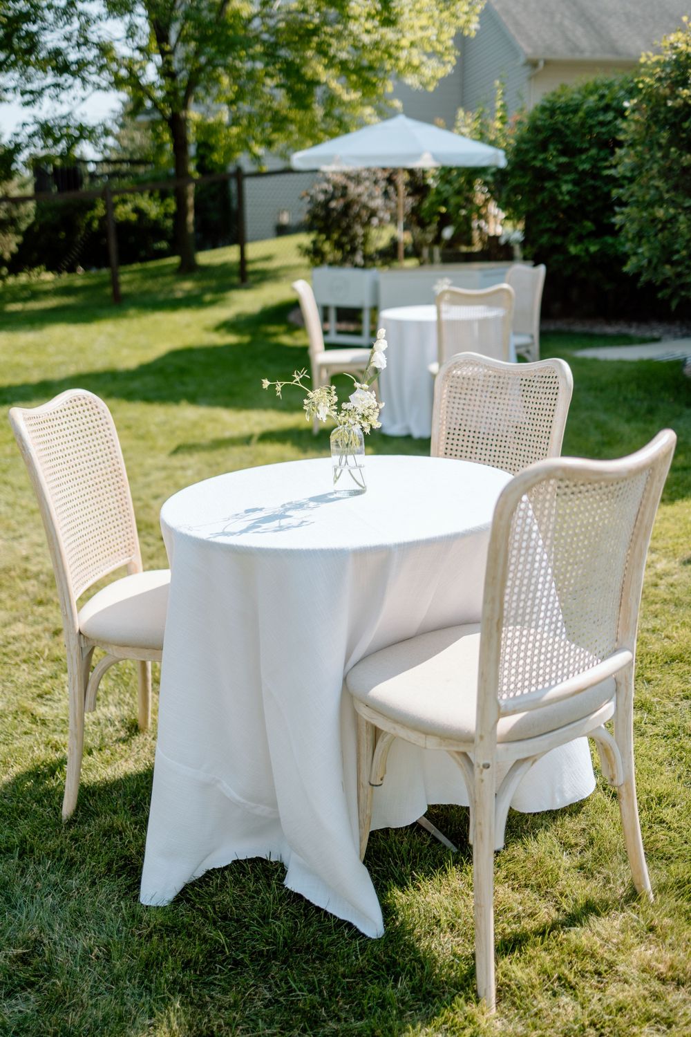 Elegant outdoor wedding setup with round table draped in white linen and vintage cane back chairs on a sunny lawn.