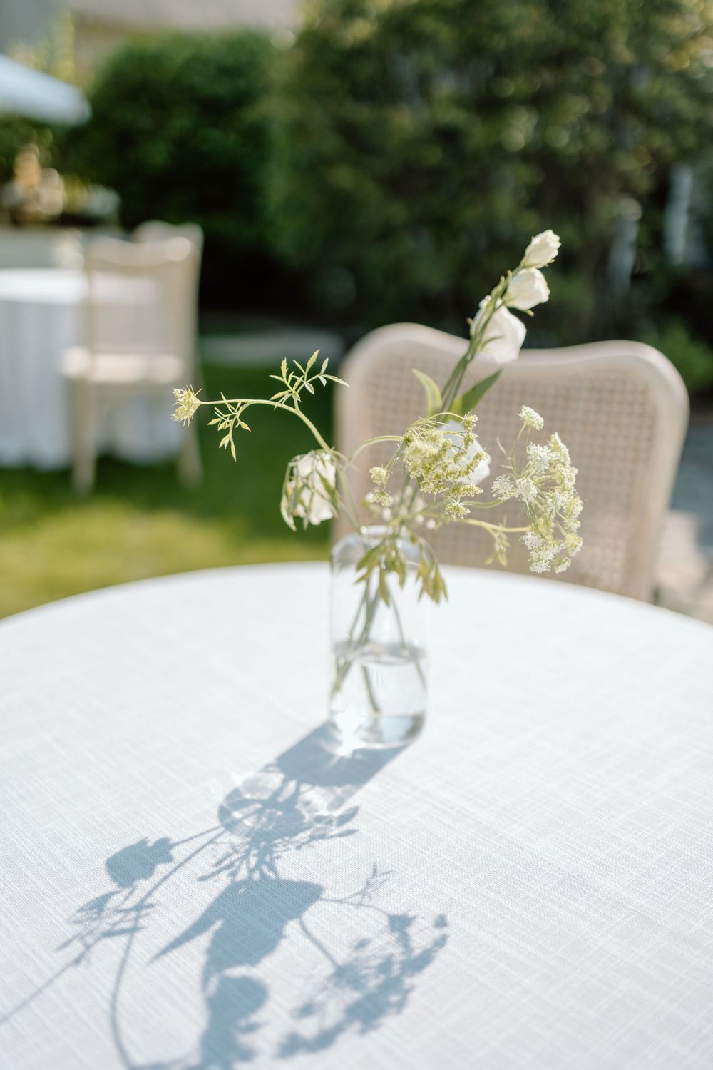 Delicate white wildflowers in a glass vase cast shadows on a white tablecloth at an outdoor garden setting.