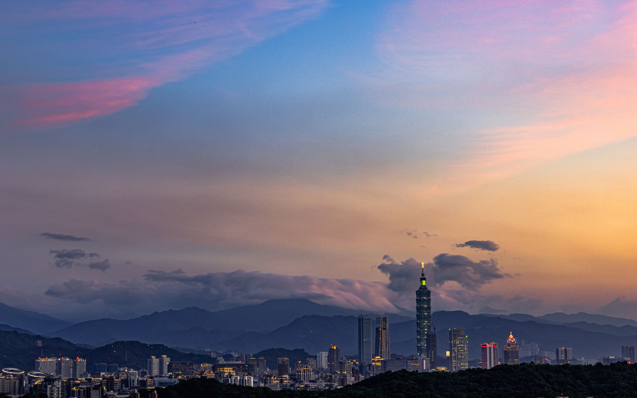 Dramatic sunset cityscape panorama with purple and orange sky over mountain silhouettes.