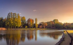 Serene autumn lake reflection with golden trees and walking path during sunset.