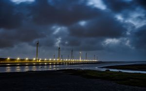 Illuminated pier stretches into dark ocean under moody storm clouds at night with glowing lights reflecting on water.