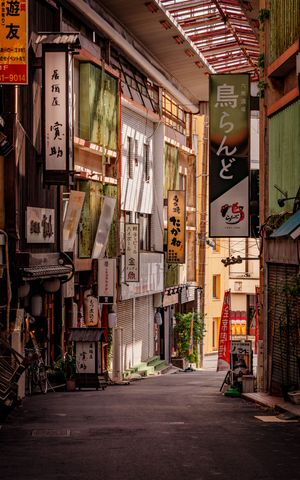 A narrow alley in Japan with traditional shops and lanterns displaying Japanese text and signs in warm ambient lighting.