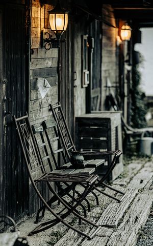 Weathered wooden folding chairs on rustic porch with glowing wall lanterns casting warm light.