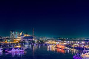 Panoramic night view of a harbor with illuminated boats and city lights reflecting on water.