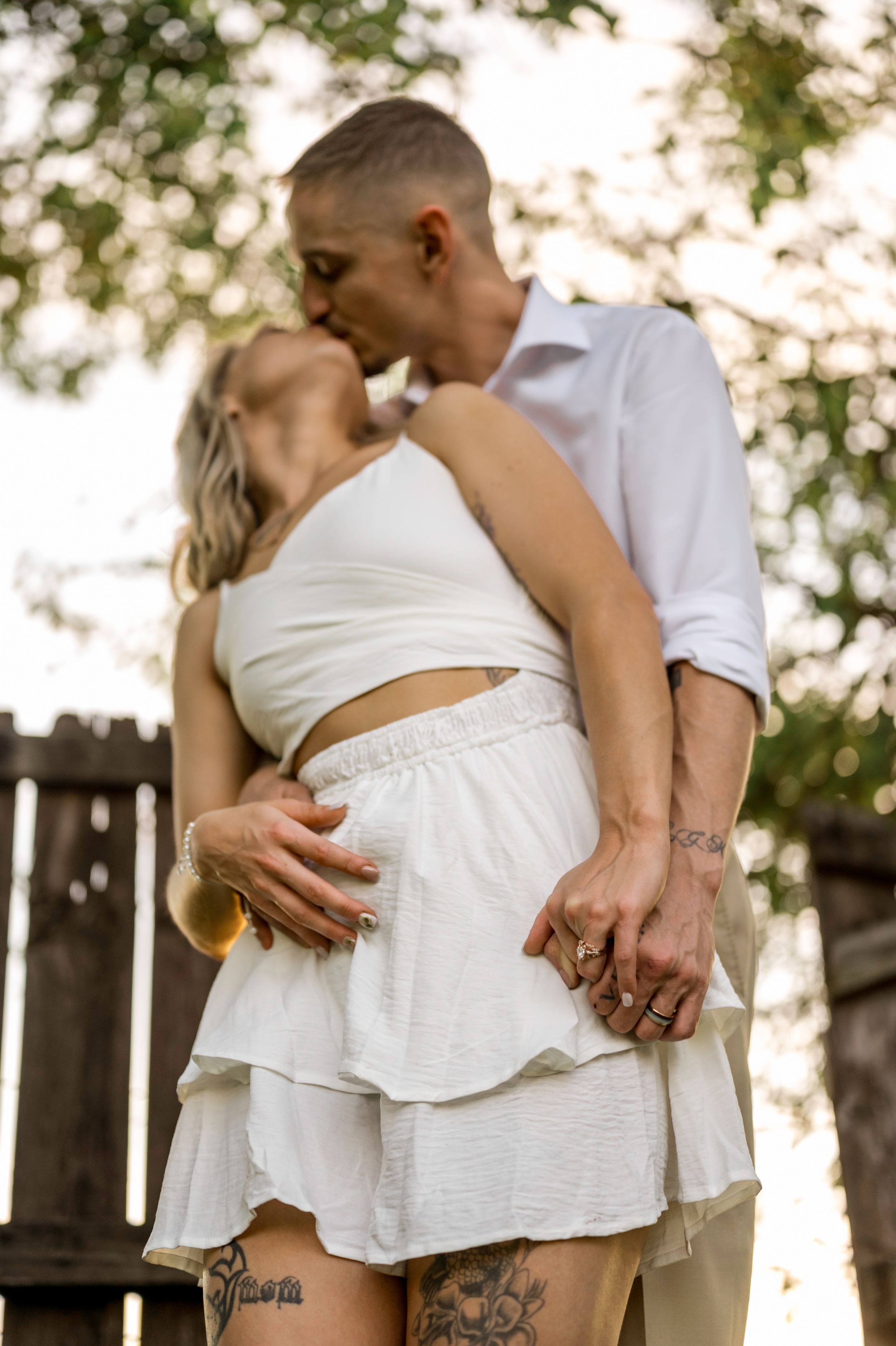 A romantic outdoor portrait of a couple embracing in white summer outfits during golden hour with visible tattoos.